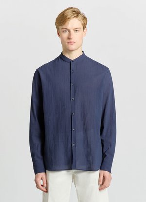 Jeune homme aux cheveux blonds portant une chemise texturée bleu marine à boutons avec col mao et un pantalon blanc, debout devant un fond uni.