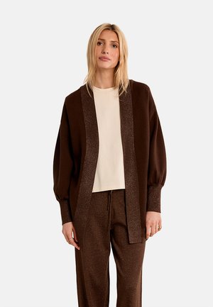 Donna bionda che indossa un cardigan marrone aperto davanti, pantaloni marroni abbinati e una maglietta beige chiaro, in piedi davanti a uno sfondo neutro.