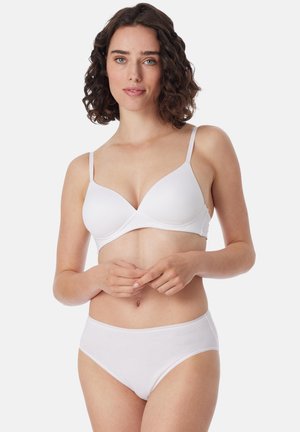 Woman with short curly hair wearing a white bra and matching white underwear, standing with hands gently clasped.