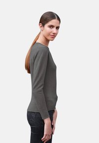 Haut gris à manches longues avec un col rond, au design ajusté et à la texture douce, associé à un jean foncé. Le modèle pose de profil.