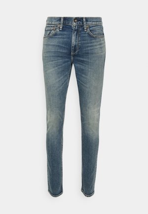 Siniset slim fit -farkut, joissa haalistuneita yksityiskohtia, etutaskut, vyölenkit ja napillinen kiinnitys yksivärisellä taustalla.