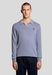 Νέος άνδρας όρθιος, φοράει ένα ανοιχτό μοβ πουλόβερ πόλο με μακριά μανίκια και λογότυπο κίτρινου πουλιού στο στήθος, καθώς και σκούρο παντελόνι.