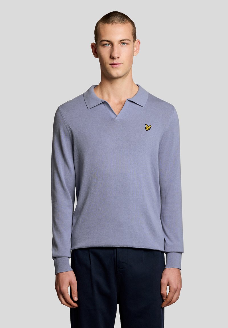 Νέος άνδρας όρθιος, φοράει ένα ανοιχτό μοβ πουλόβερ πόλο με μακριά μανίκια και λογότυπο κίτρινου πουλιού στο στήθος, καθώς και σκούρο παντελόνι.