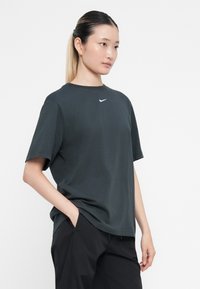 Tumši pelēks īsās piedurknes t-krekls ar apaļu kakla izgriezumu, uz krūtīm redzams balts Nike logo. Audums izskatās mīksts un gluds.