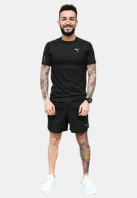 Camiseta deportiva negra de manga corta, con un bolsillo en la parte delantera. Combinada con pantalones cortos negros y zapatillas blancas. Tatuajes coloridos notables en la pierna.