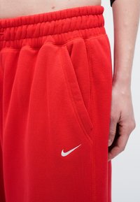Crvene trenirke s elastičnom pojasevima, bočnim džepovima i suptilnim bijelim Nike logom. Materijal izgleda mekano s glatkom teksturom.