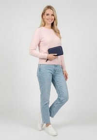 Smiling woman in pink sweater, light blue jeans, and white sneakers stands holding a navy blue zip wallet against a plain background.