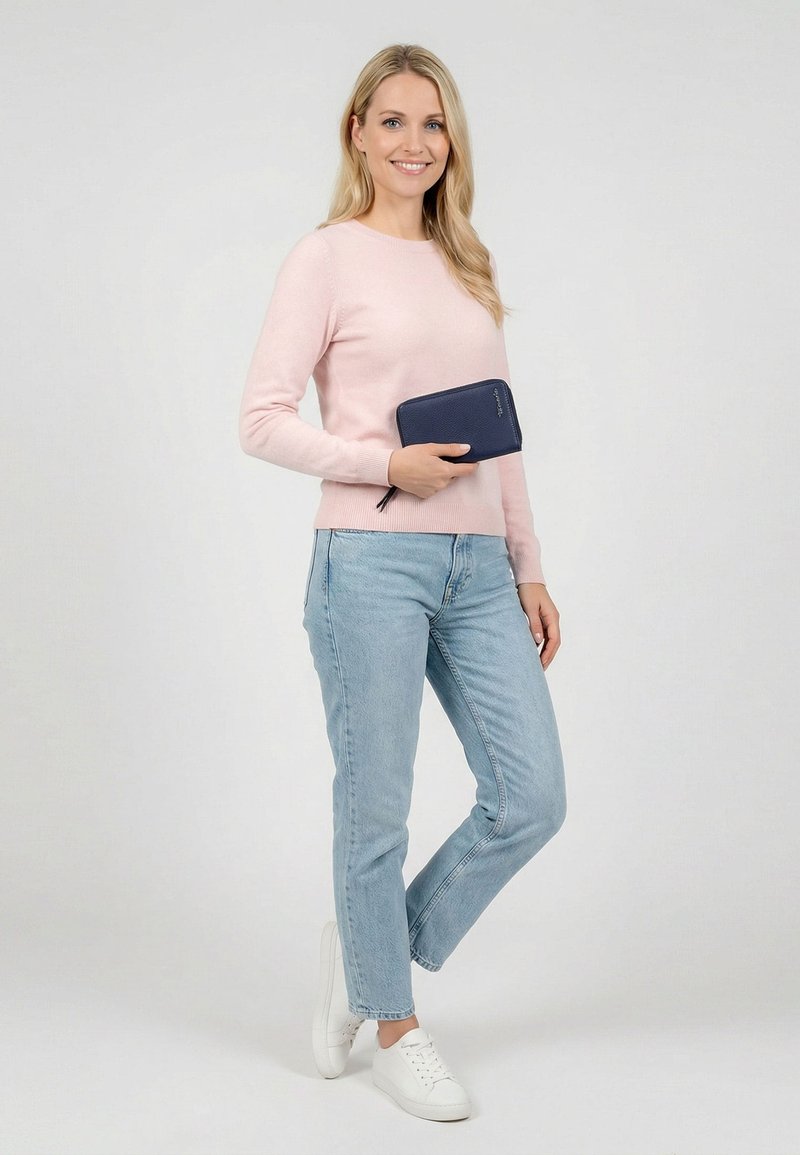 Smiling woman in pink sweater, light blue jeans, and white sneakers stands holding a navy blue zip wallet against a plain background.