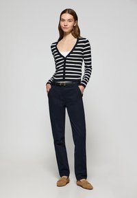 Randig marinblå och vit kofta med V-ringning, tillsammans med mörka byxor. Modellen står med händerna i fickorna, iförd beige loafers.