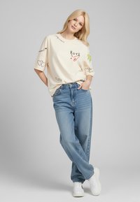 T-shirt crème surdimensionné avec divers imprimés graphiques, y compris des cœurs et le mot "amour", associé à un jean baggy bleu et des baskets blanches.