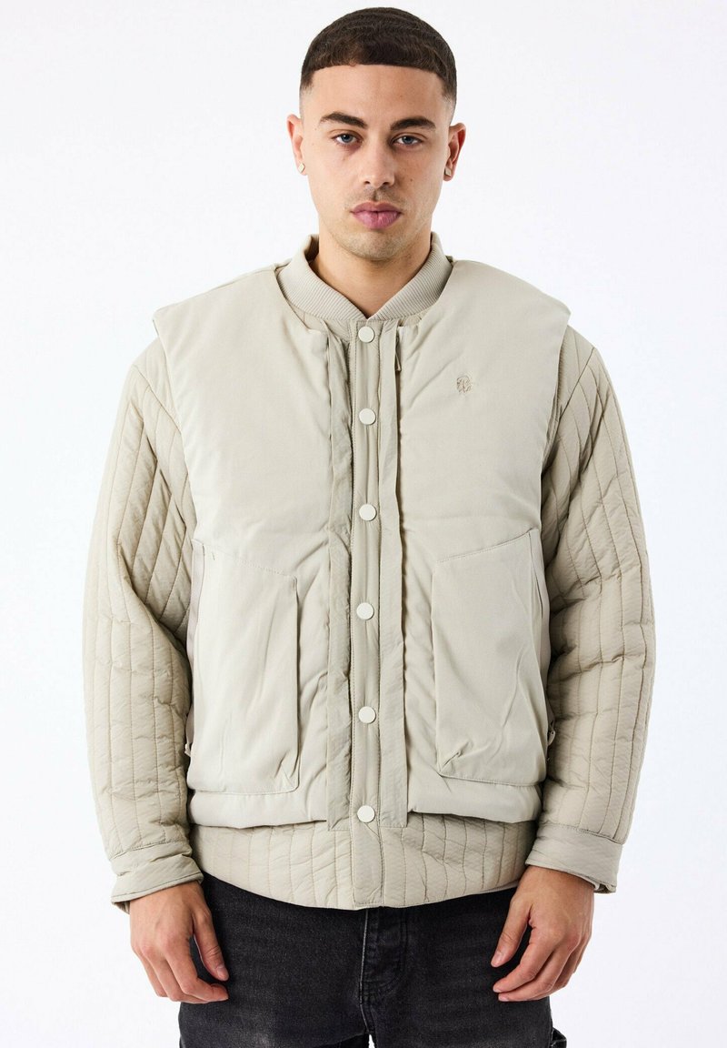 Veste matelassée beige avec un col côtelé, fermeture à boutons-pression à l'avant et deux poches avant. Présente un extérieur lisse et des manches texturées.