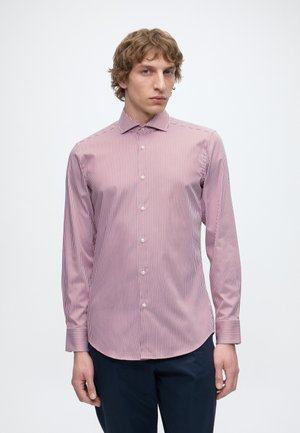 Homme aux cheveux bouclés portant une chemise à rayures verticales roses et blanches avec boutons et un pantalon foncé, debout devant un fond clair uni.