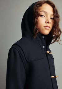 Ung person med lockigt hår iförd en svart huva rock med träknappar, som ser något åt sidan mot en neutral bakgrund.