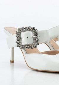 Chaussures à talons hauts blanches avec une large bride ornée d'une boucle en strass argenté. Finition en cuir lisse et design à bout pointu.