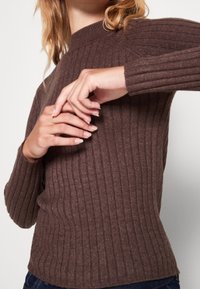 Bruin geribbeld gebreid sweater met een hoge hals, lange mouwen en een aansluitende pasvorm. De textuur lijkt zacht en knus.