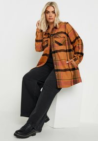 Rutig skjorta i orange, bruna och svarta toner; har knappar och två bröstfickor. Bärs över svarta vida jeans och svarta skor.