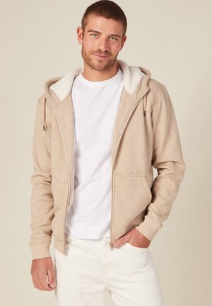 Beige zip-up hoodie with a soft white fleece-lined hood, featuring front pockets and ribbed cuffs, worn over a white t-shirt and light pants.