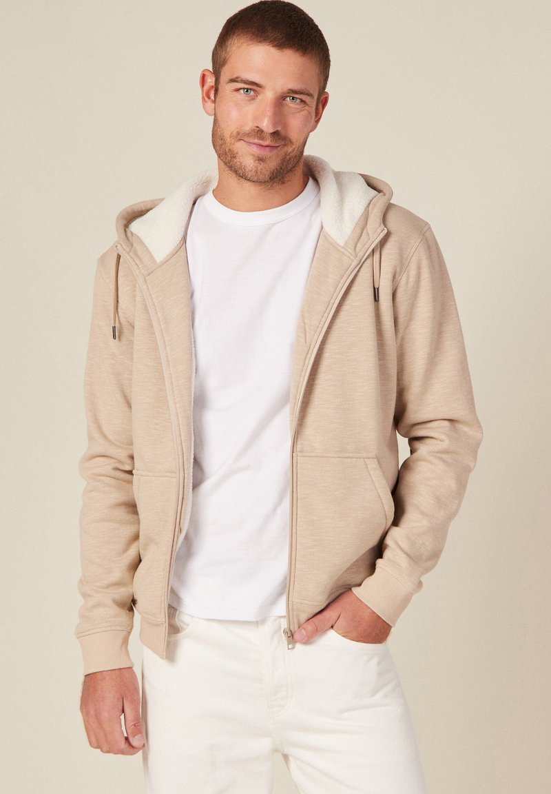 Felpa beige con zip e cappuccio foderato in morbido pile bianco, con tasche frontali e polsini a coste, indossata su una t-shirt bianca e pantaloni chiari.