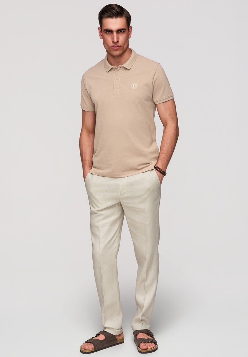 Homme debout, les mains dans les poches, portant un polo beige, un pantalon clair et des sandales marron foncé sur un fond uni.
