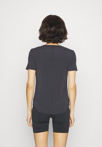 T-shirt nera a maniche corte con orlo arrotondato, realizzata in un tessuto morbido e liscio. Indossata con pantaloni corti neri aderenti, mostrata da dietro.