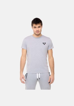 Giovane uomo che indossa una maglietta aderente grigio chiaro con un piccolo logo sul petto sinistro e pantaloni della tuta grigi abbinati con tasche con zip nere.