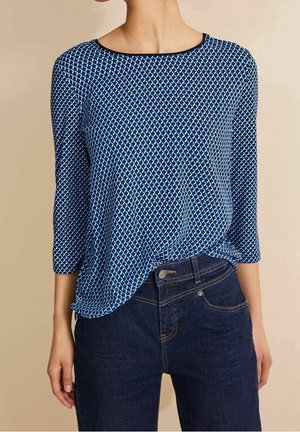 Blouse bleue à motifs avec manches trois-quarts et col rond, en tissu léger. Portée avec un jean en denim foncé.