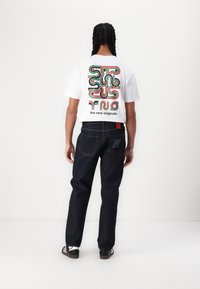 White t-shirt with colorful abstract graphic on the back, paired with dark denim jeans featuring subtle branding on the pocket.