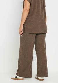 Bruine geribbelde gebreide top met zijsplitten in combinatie met bijpassende wijde broek; heeft een zachte textuur en een relaxed fit, aangevuld met witte sneakers.