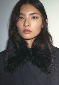 Sort faux pelskrave med lange fibre, sat mod et mørkt tøj. Den glatte, skinnende tekstur kontrasterer med kravens fylde.