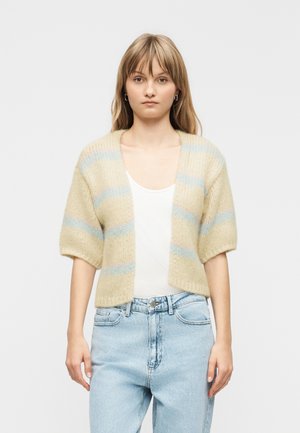 Jeune femme debout, face à l'avant, portant un cardigan à manches courtes rayé beige et bleu clair, un haut blanc et un jean bleu clair sur un fond uni.