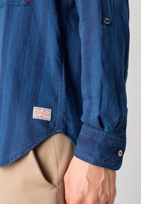 Cămașă din denim albastru cu dungi verticale, având un eticheta cusută la tiv și manșete cu nasturi. Textură netedă și croială lejeră.
