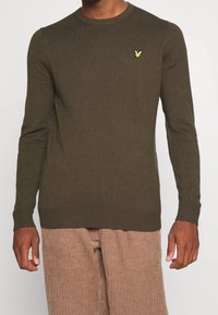 Pull tricoté vert olive avec un col rond et des poignets côtelés. Présente un petit logo jaune clair sur la poitrine.