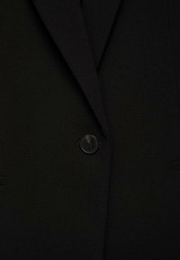 Blazer noir avec une texture de tissu lisse, des revers en pointe et une fermeture à un bouton noir. Comprend deux poches latérales.