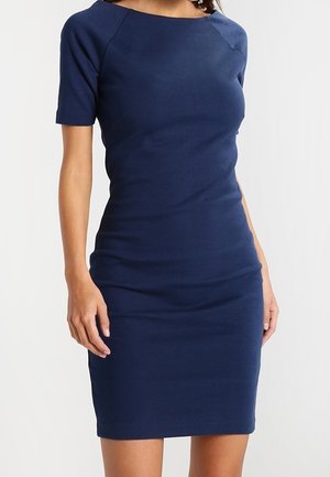 Mujer que lleva un vestido azul marino ajustado hasta la rodilla con mangas cortas y escote barco, de pie frente a un fondo claro y sencillo.