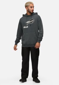 Sudadera gris con el texto "BILLS" y el logo en el frente, bolsillo tipo canguro y cordones, combinada con pantalones cargo negros y zapatos negros.