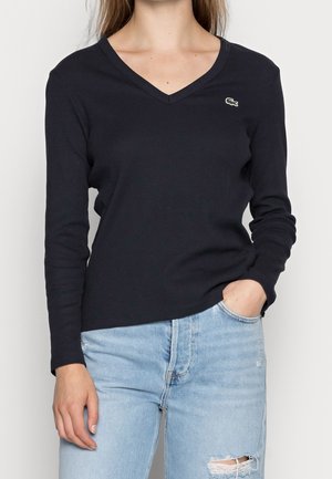 Femme portant un haut noir à manches longues et col en V avec un petit logo crocodile et un jean bleu clair déchiré, une main dans la poche.