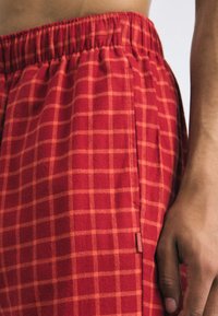 Red checkered shorts made of soft fabric, featuring an elastic waistband and side pockets. The check pattern has lighter orange lines.