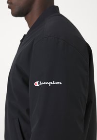 Svart nylon bomberjacka med dragkedja fram, ribbad krage och logotyp på vänster ärm i vitt och rött. Slät textur, åtsittande design.