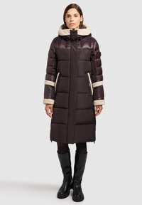 Cappotto imbottito marrone con cappuccio in pelliccia sintetica, caratterizzato da cuciture verticali e tasche con cerniera. Il cappotto presenta polsini beige a contrasto e dettagli accentuati.