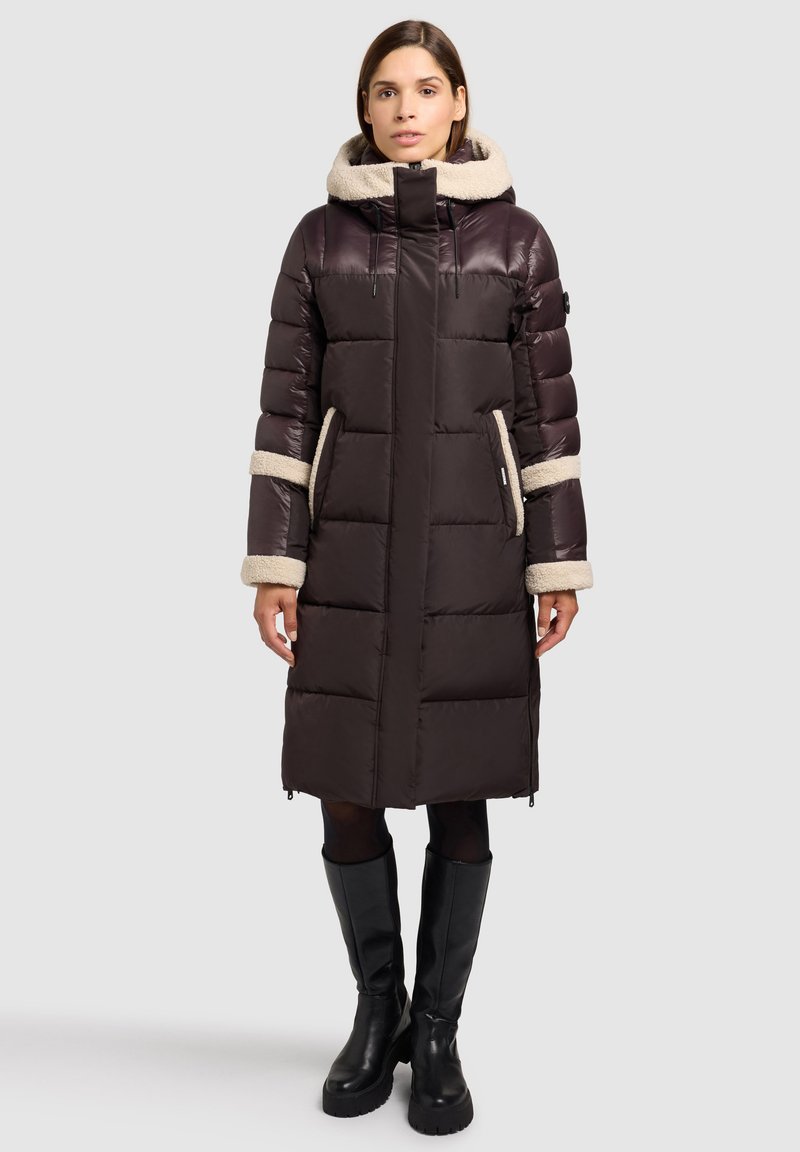 Cappotto imbottito marrone con cappuccio in pelliccia sintetica, caratterizzato da cuciture verticali e tasche con cerniera. Il cappotto presenta polsini beige a contrasto e dettagli accentuati.