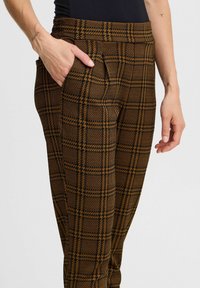 Pantalons à carreaux marron et noir avec une texture lisse, dotés de poches latérales et d'une taille ajustée. Gros plan sur une main reposant dans la poche.