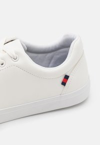 Sneaker bianco con materiale sintetico liscio, punta arrotondata e suola in gomma. L'interno presenta una fodera in tessuto grigio; dettaglio di branding rosso e blu sul lato.