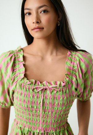 Kobieta o długich, ciemnych włosach, nosząca złote kolczyki koła oraz zielono-różową sukienkę w kratę z bufkami i marszczonym stanem.