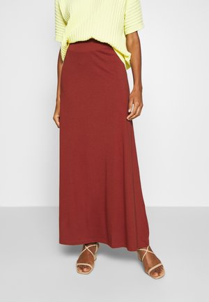 Femme portant une longue jupe couleur rouille, un pull à manches courtes jaune pâle et des sandales beiges à lanières, debout devant un fond uni.
