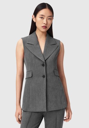 Vrouw draagt een mouwloos grijs op maat gemaakt colbert met inkeping revers, knoopsluiting en bijpassende grijze broek tegen een effen achtergrond.