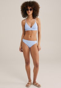 Ljusblå och vit randig bikiniuppsättning med triangeltopp och medeldäckande botten. Modellen bär runda solglasögon och sandaler.