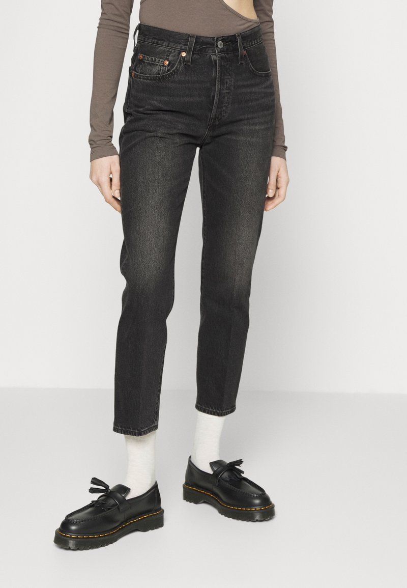 Personne portant un jean noir taille haute, des chaussettes blanches, des mocassins noirs à pampilles et un haut marron ajusté à manches longues, debout sur un sol clair.