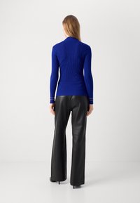 Femme aux cheveux longs portant un pull côtelé bleu ajusté et un pantalon large en cuir noir, debout de dos sur un fond blanc uni.