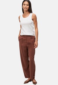 Top bianco senza maniche con un motivo tessurizzato abbinato a pantaloni larghi marroni. La modella indossa delle scarpe flats marroni. Sfondo semplice.