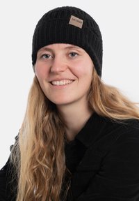 Czarna dzianinowa czapka z teksturowanym wzorem i małą brązową skórzaną metką. Osoba ma długie, falujące blond włosy i nosi czarną kurtkę.